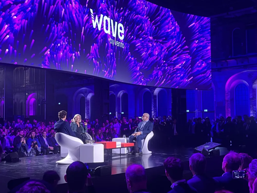 Jony Ive en John Elkann in gesprek met Zanny Minton Beddoes in witte stoelen aan een rode tafel, onder het Wave-logo.