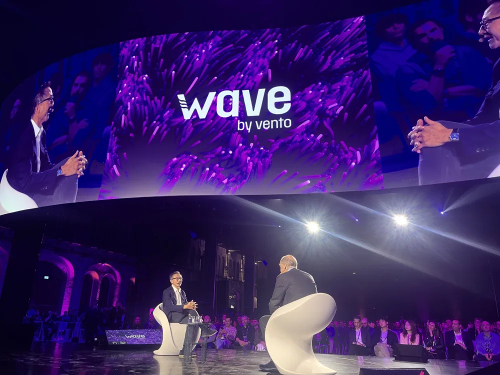 Luca Bocchio en Luca De Angelis in gesprek op witte stoelen op het hoofdpodium van Wave by Vento, met hun beeld groot op het scherm erachter.